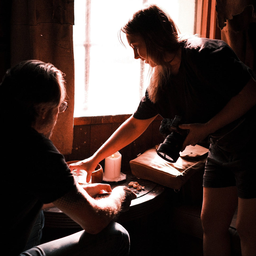 A dimly lit indoor scene with two people gathered around a table near a bright window. One person stands and holds a camera, while the other sits and appears to be interacting with items on the table.