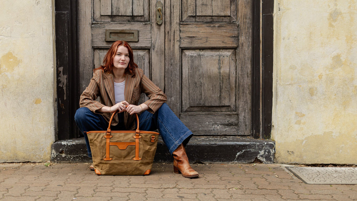 A woman with red hair sits on the steps of a rustic wooden door, holding a large tan canvas tote bag with orange-brown leather handles and trim. She is wearing a brown jacket, blue jeans, and brown leather boots.